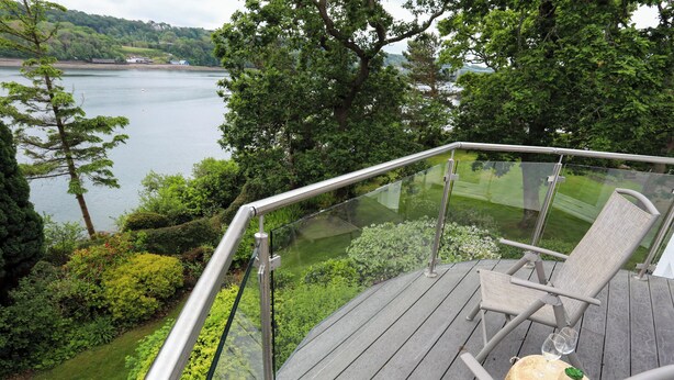 A deck with glass railings overlooking a lake, equipped with two lounge chairs and a small table with a glass of wine. - Ynys Castell (Menai Bridge)