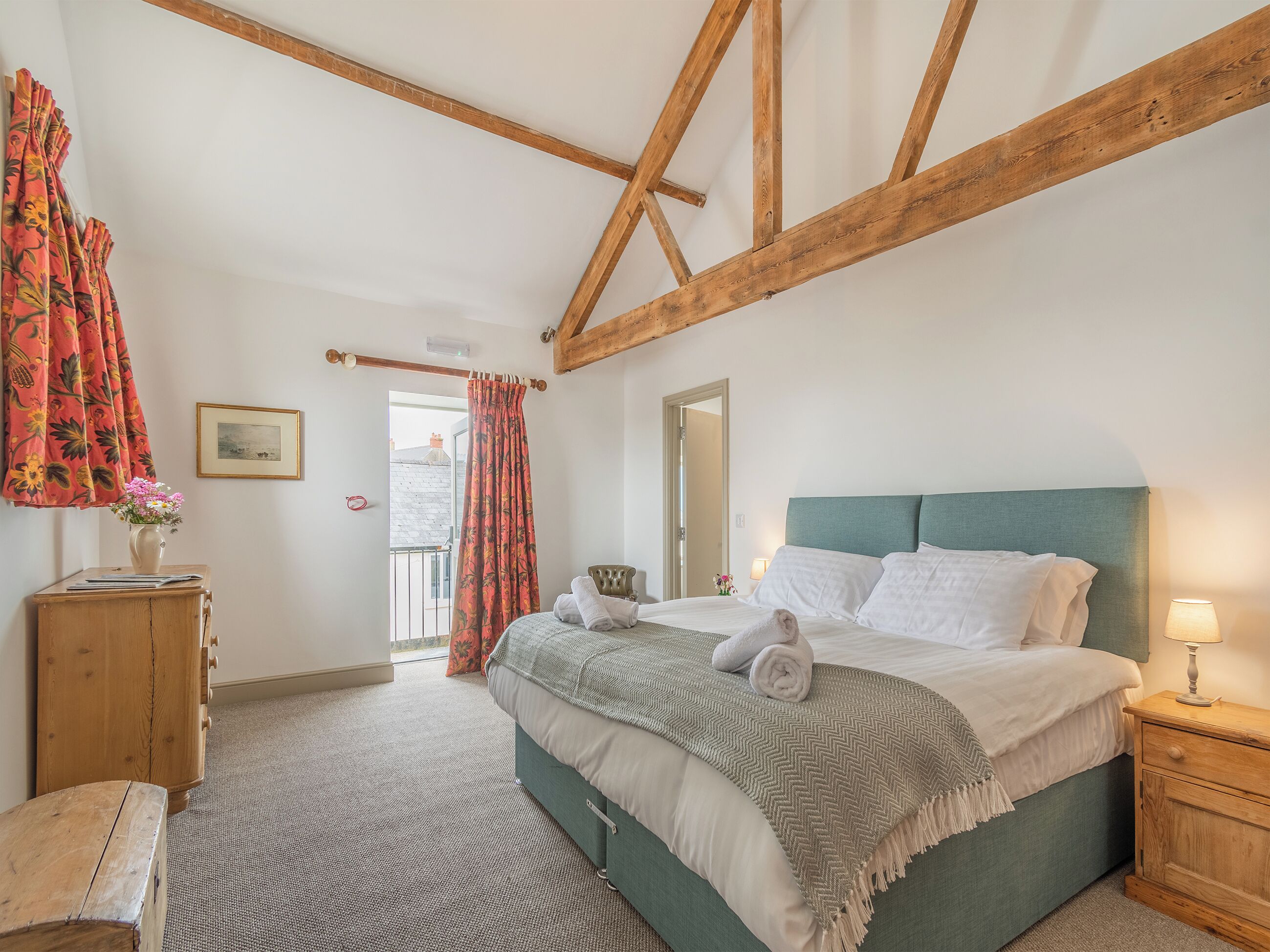 Une chambre à coucher avec un lit, une tête de lit en bois, une table de chevet, une commode en bois, un tableau encadré au mur et une fenêtre avec des rideaux.