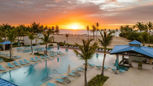 A resort pool area with lounge chairs and umbrellas, a beach in the background, and a sunset.