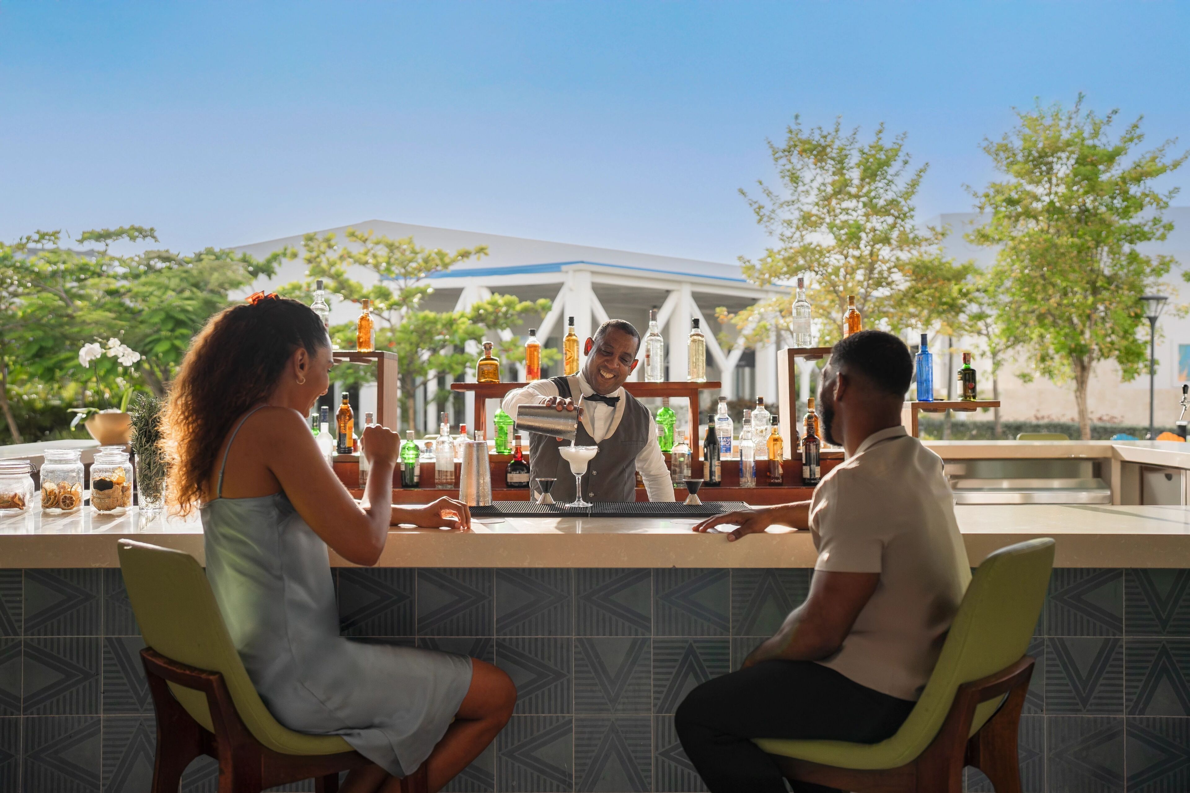 A bar area with a bartender preparing drinks, a counter with various bottles, and two seated guests.