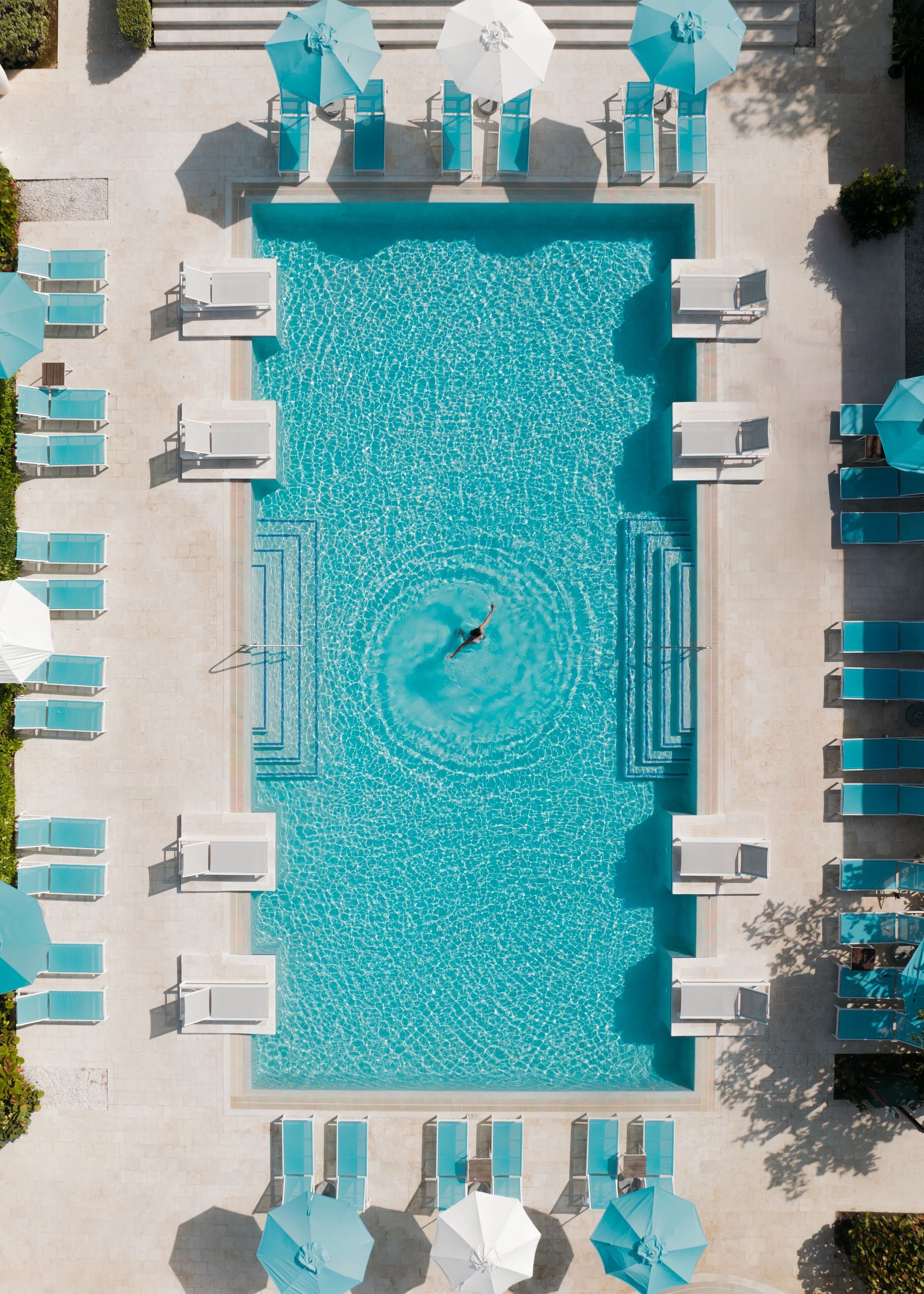 An aerial view of a rectangular swimming pool with a person creating a circular wake pattern. Surrounding the pool are multiple lounge chairs and umbrellas, arranged in rows.