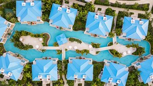 An aerial view of a resort with multiple blue-roofed villas arranged around a central swimming pool, surrounded by lush greenery and palm trees.