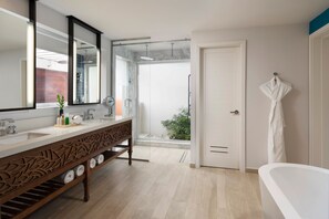 A modern bathroom with a large mirror, two sinks, a bathtub, and a door leading to a balcony with plants.