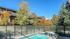 Un bain à remous rectangulaire, entouré de pierres, situé dans un jardin avec une clôture en métal et des arbres aux feuilles d’automne.