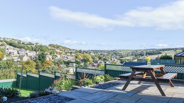 Um pátio com mesa e bancos de madeira, cercado por uma vedação verde e canteiros de jardim, com vista para uma área residencial com casas e colinas.