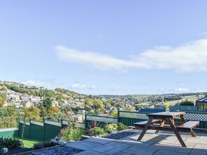 A patio with a wooden table and benches, surrounded by a green fence and garden beds, overlooking a residential area with houses and hills. - Holme Valley Cottage (Holmfirth)