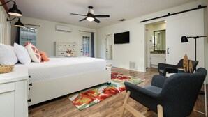 Quarto com cama, cadeira, ventoinha no teto, televisão e porta para um banheiro.