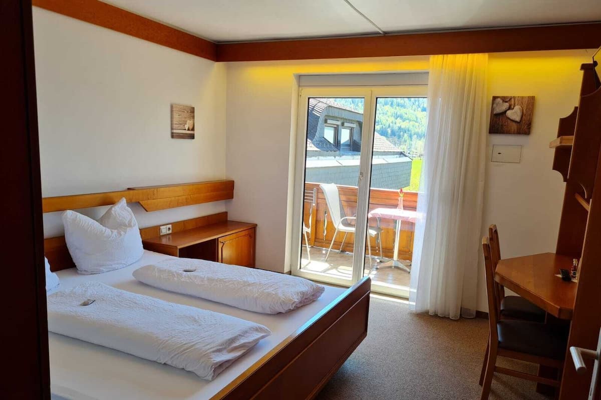 A hotel room with a large bed, a desk, a chair, and a view of a balcony with outdoor seating.