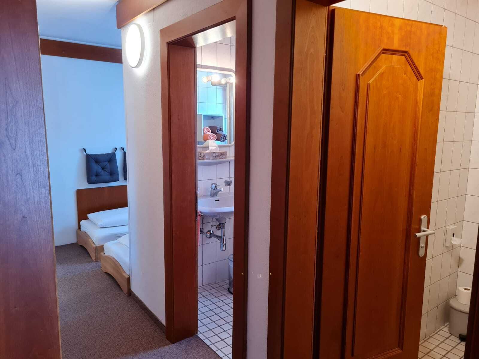 Habitación de hotel con puerta de madera, cama con ropa de cama blanca, mesita de noche y un pequeño cojín azul.