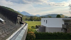 Blick aus einem Fenster auf eine grüne Wiese, ein modernes Gebäude und ferne Berge.
