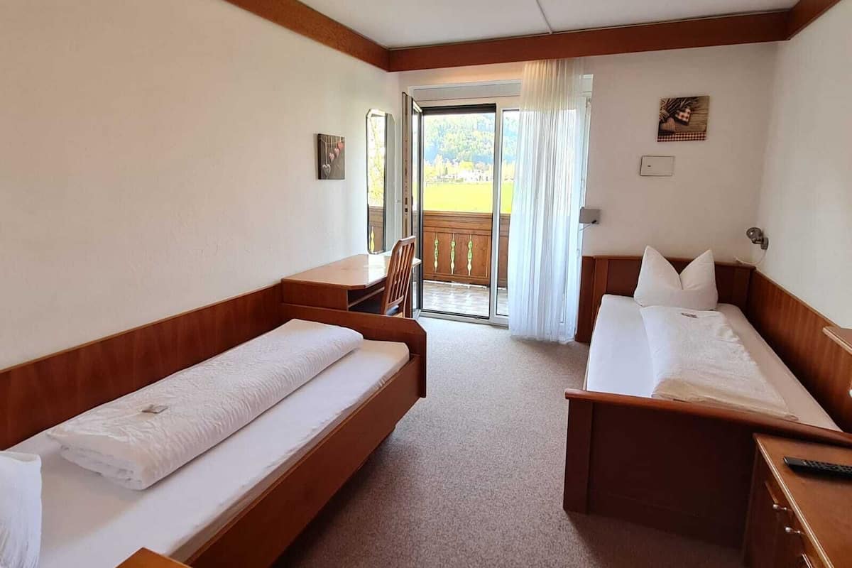 A hotel room with two single beds, a wooden desk, a chair, and a balcony with a view.