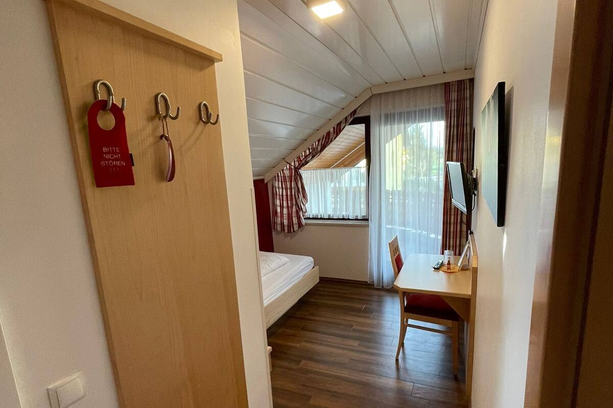 A room with a bed, a desk, and a TV mounted on the wall. There is a window with curtains and a door with a coat rack.