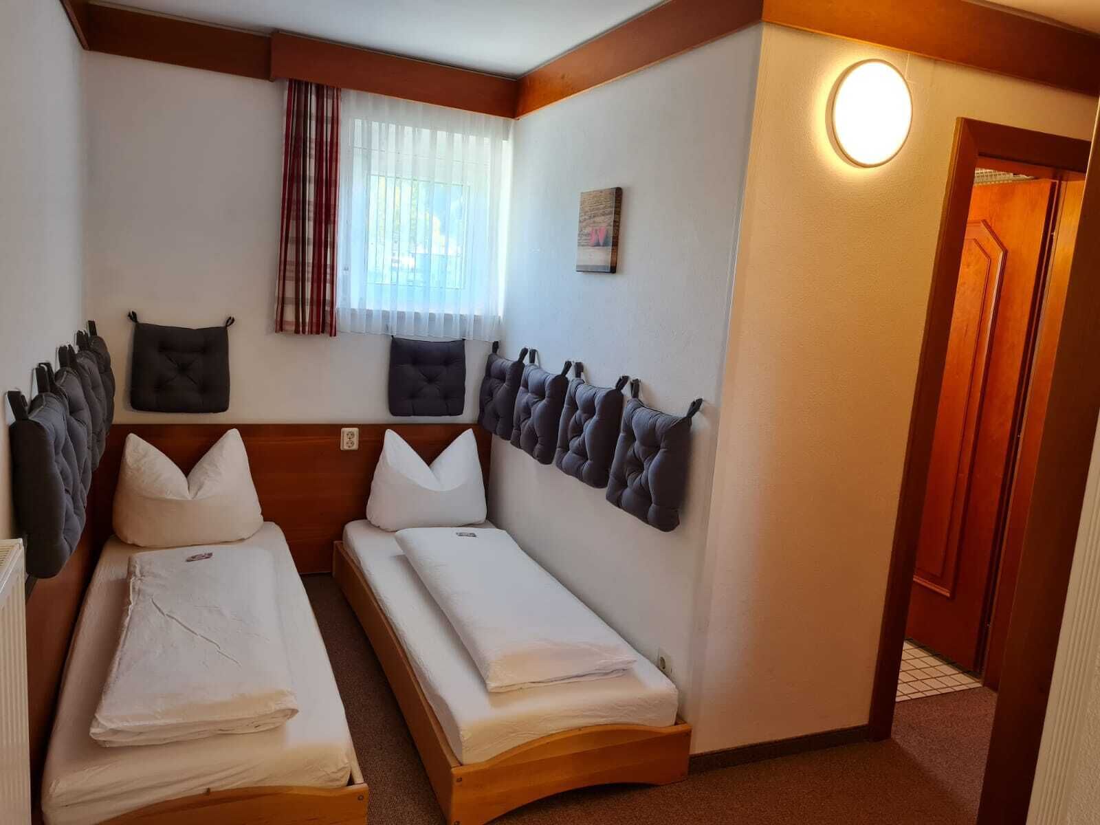 A hotel room with two single beds, a wooden headboard, a window with curtains, a wall-mounted light, and a door.
