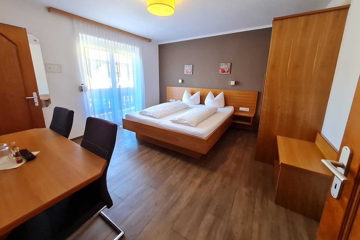 A hotel room with a wooden bed, a desk with chairs, a large window with curtains, and a ceiling light.
