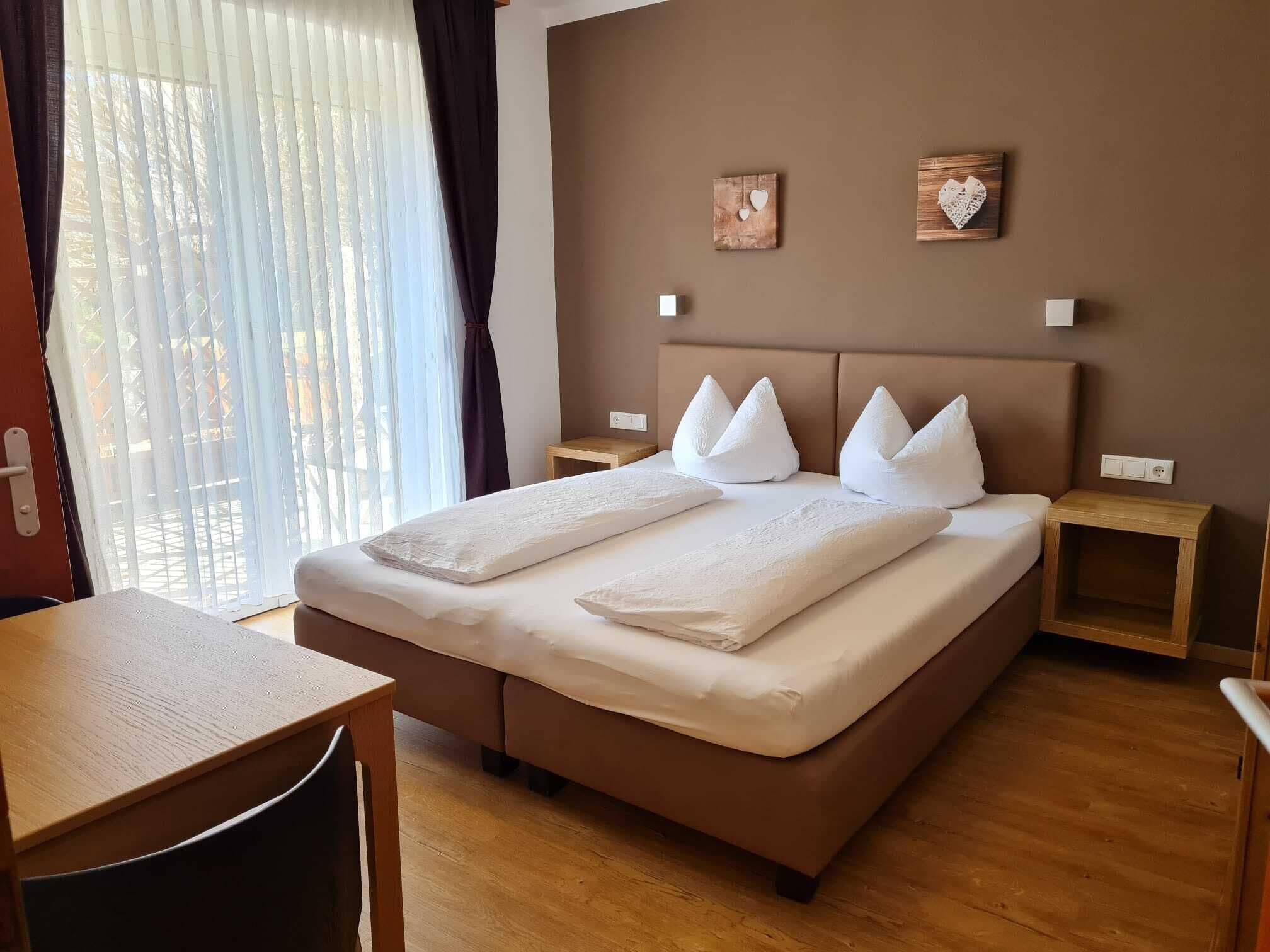 Ein ordentlich eingerichtetes Hotelzimmer mit einem großen Bett, zwei Nachttischen, einem kleinen Holztisch und Blick durch ein Fenster mit transparenten Vorhängen.