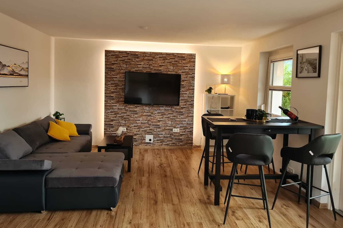 A modern living room with a grey sofa, a flat-screen TV mounted on a stone wall, a dining area with a table and chairs, and a window with a view of greenery.