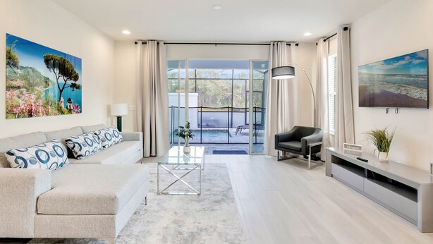 Een moderne woonkamer met een bank, salontafel en tv-meubel. Er is een groot raam met gordijnen, een plant en uitzicht op een zwembad en terrasmeubilair.