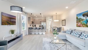 Een moderne woonkamer met een grijze bank, een glazen salontafel en een keuken op de achtergrond.