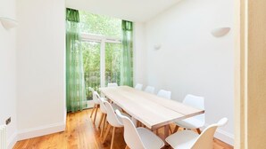 A modern dining room with a long wooden table and white chairs, large windows with green curtains, and a view of greenery outside. - The Woolwich Retreat - Enticing 5bdr House With Parking (London)