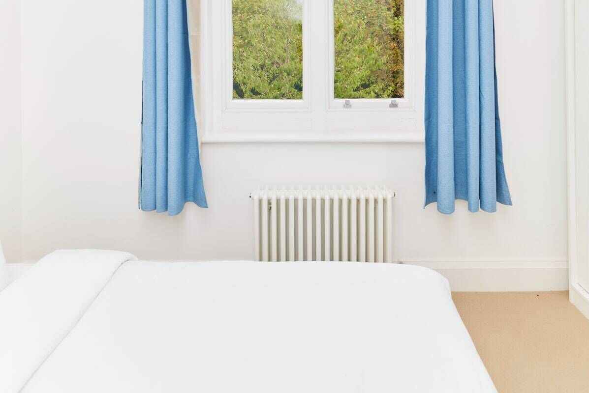 A bedroom with a bed, white walls, a window with blue curtains, and a radiator.