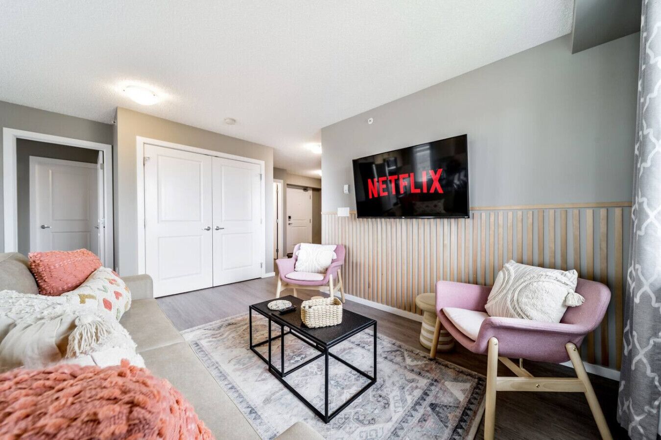 Ein Wohnzimmer mit einer Couch, zwei Sesseln, einem Couchtisch und einem an der Wand montierten Fernseher, der 'NETFLIX' anzeigt.