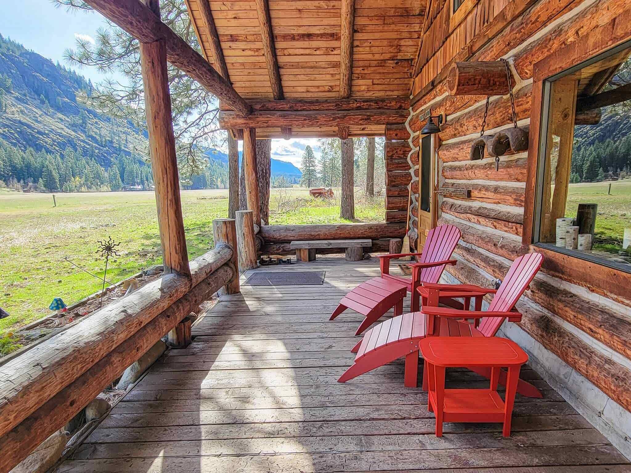 A log cabin with a wooden porch, red Adirondack chairs, and a view of a green field and mountains.