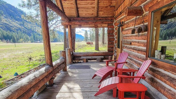 A log cabin with a wooden porch, red Adirondack chairs, and a view of a green field and mountains. - Goat Wall Cabin: log cabin retreat on the meadow (Mazama)