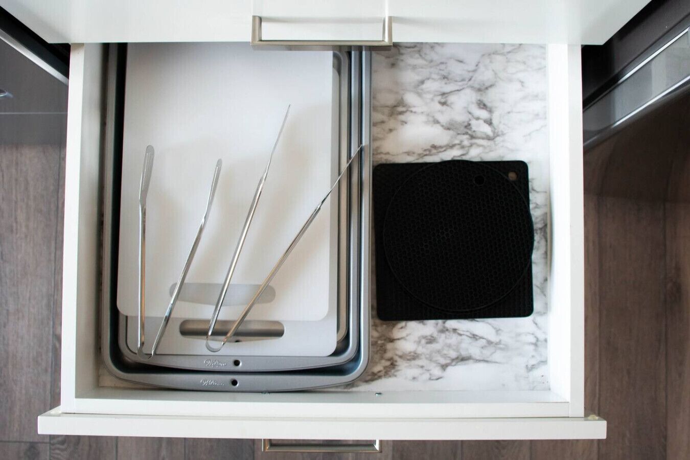 A kitchen drawer with a marble countertop, containing a black cutting board, a metal tray, and three metal utensils.