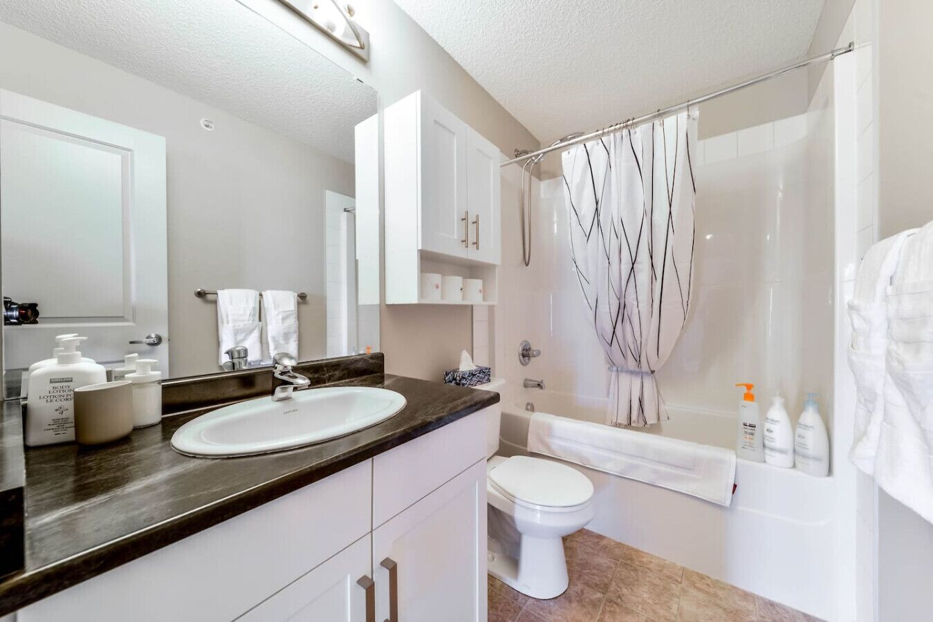 A modern bathroom with a white sink, vanity, and toilet. There are toiletries on the counter and a shower curtain with a tree design.