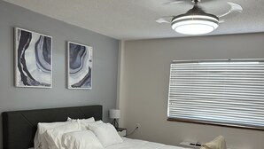 Una camera da letto con un letto, un comodino, una lampada, una ventola a soffitto e una finestra con persiane.