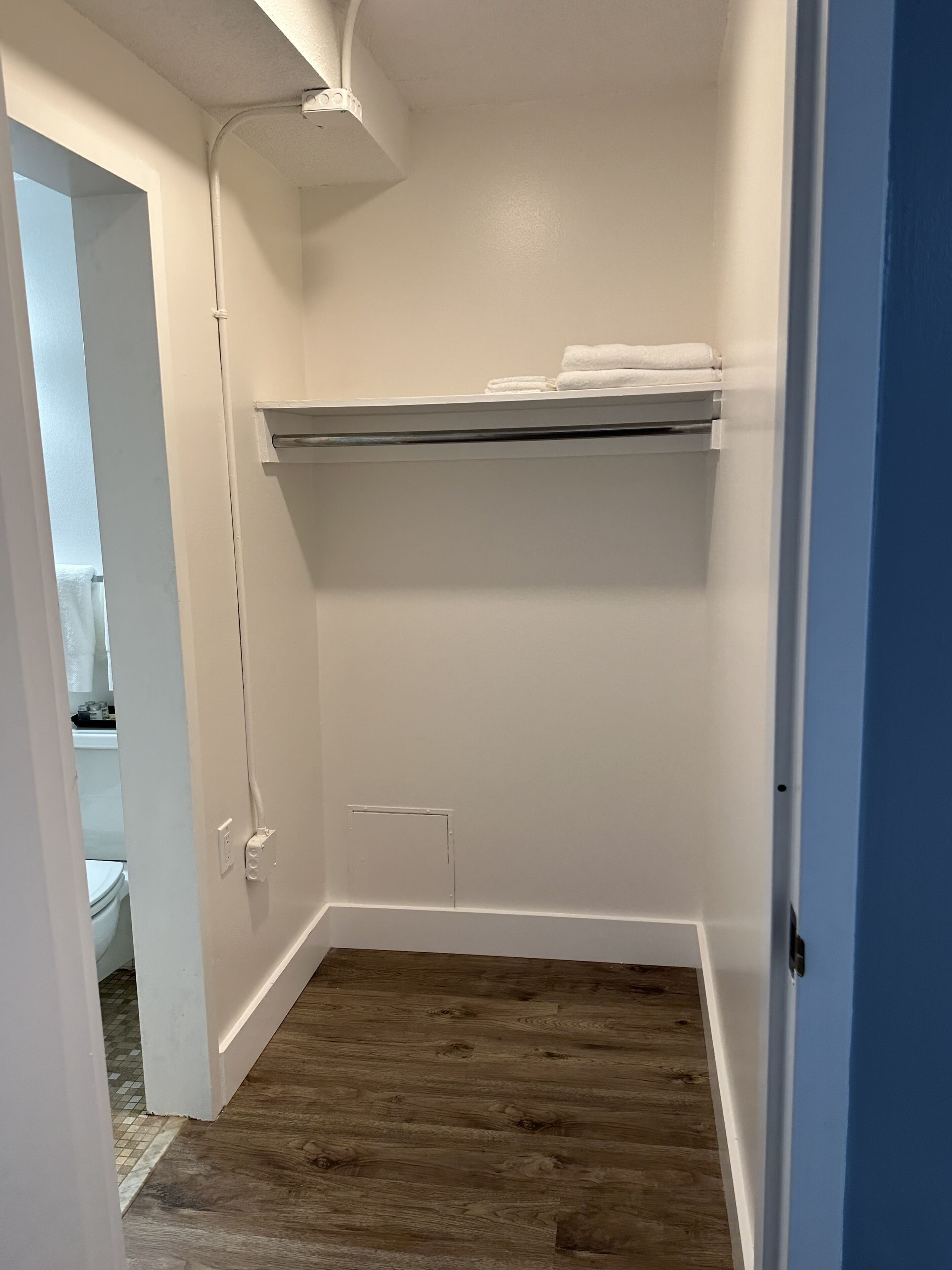 A small, white-walled closet with a wooden floor, a shelf with folded towels, and a door leading to a bathroom.