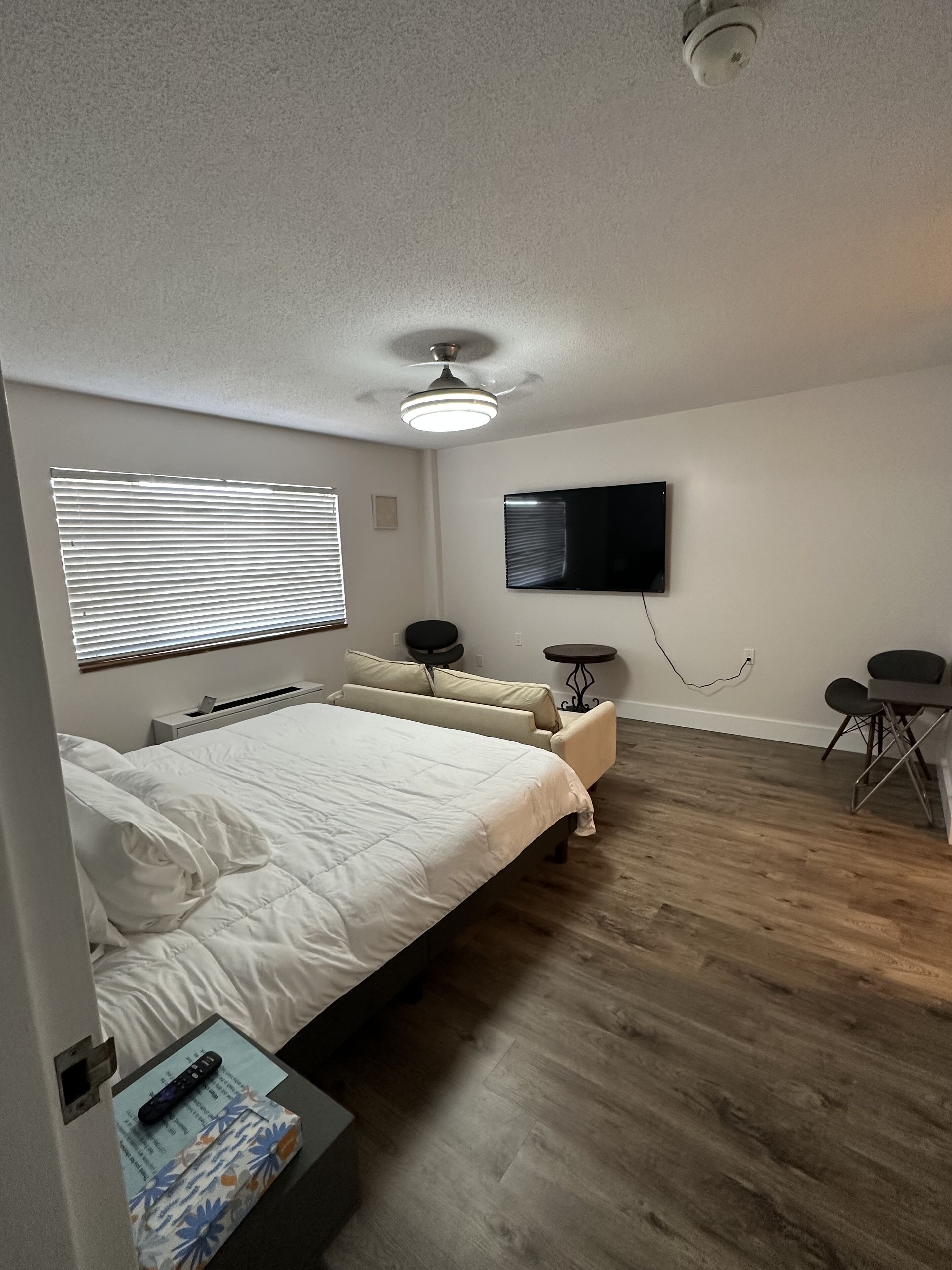 A bedroom with a bed, a ceiling fan, a TV, a small table, and a window with blinds.