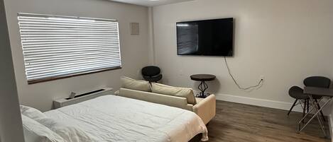A bedroom with a bed, a ceiling fan, a TV, a small table, and a window with blinds.