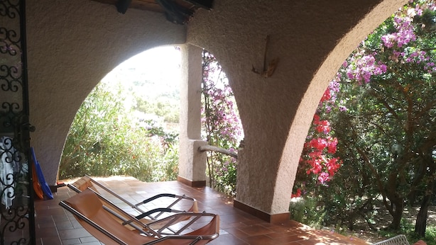 A covered patio with a wooden bench and a metal chair, surrounded by arches and a garden with flowering plants.