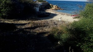A coastal path with rocky terrain and sparse vegetation, leading to a beach with boats and a clear view of the sea.