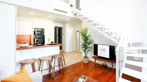 A modern kitchen with white cabinets, wooden stools, and a central island. A dining area with a white table and chairs is visible. A staircase leads to an upper level. - Young Spirit Apartment (Faro)