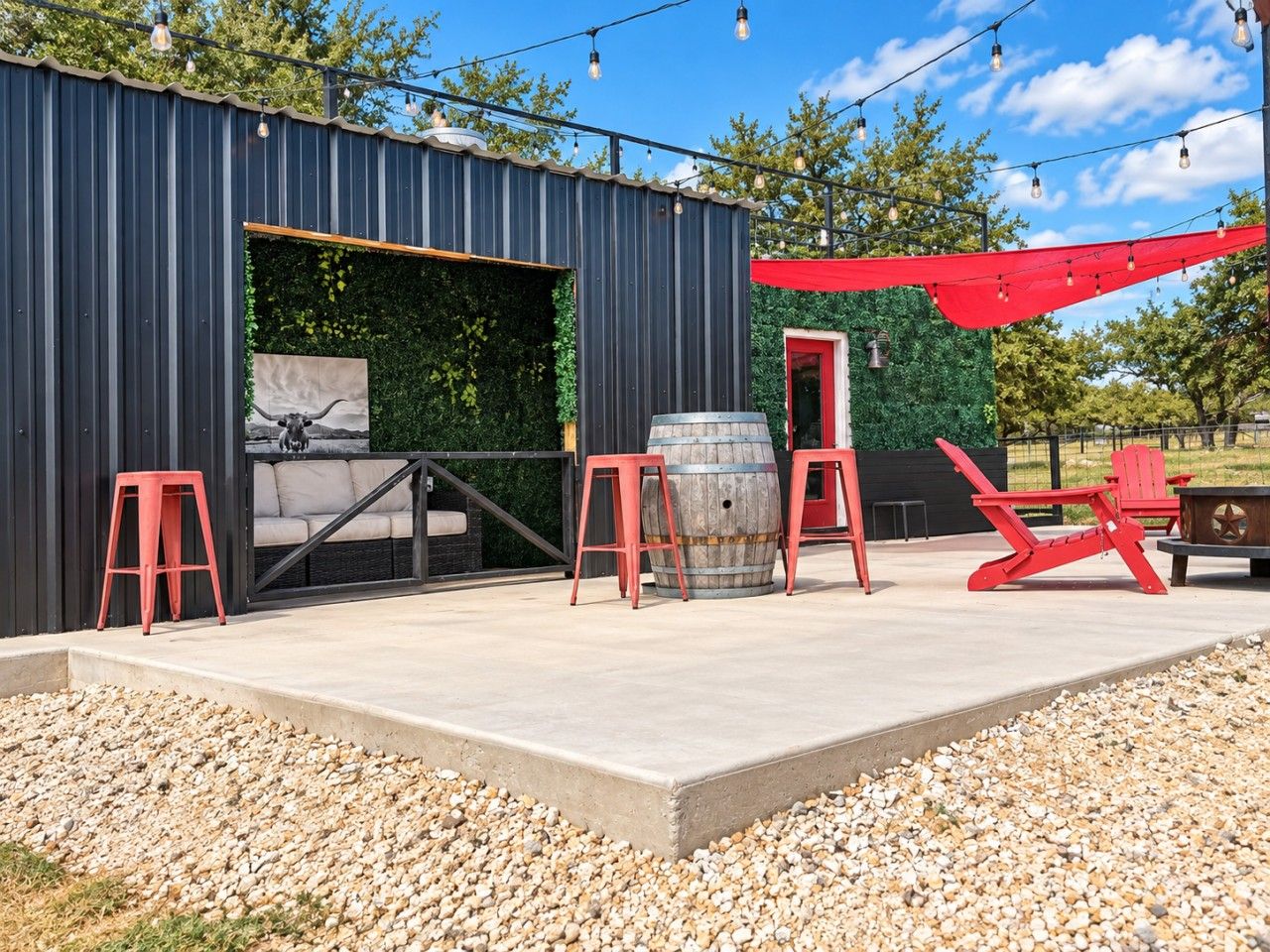 Une terrasse extérieure dotée d’un sol en béton, entourée de galets, avec une structure en métal noir, des chaises rouges, un tonneau en bois et un auvent rouge.