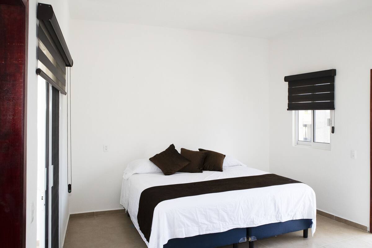 Ein Schlafzimmer mit Bett, Fenster mit Jalousien, einer Tür und einem an der Wand befestigten Regal.
