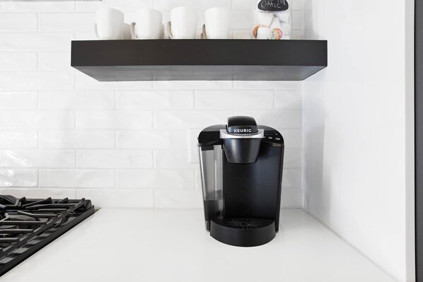 Une cuisine moderne avec une machine à café noire, un pare-feu blanc en carreaux et une étagère noire au-dessus, sur laquelle sont posées des tasses blanches et une dosette de café.