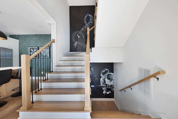 Un escalier moderne avec des marches en bois et des rampes en métal, menant à un palier orné d’une fresque en noir et blanc représentant un musicien.