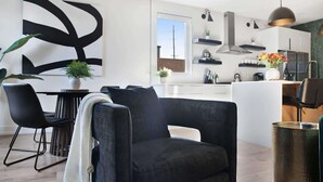 Una sala de estar moderna con un sofá negro, un comedor con mesa y sillas, y una cocina con armarios blancos y un ventanal grande.