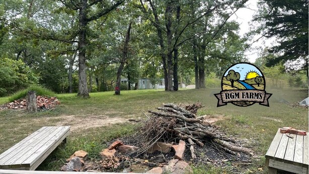 A campsite with a fire pit surrounded by wooden benches, trees, and a logo for RGM Farms. - Primitive Campsites with water spigot, toilets, and wifi! (Bourbon)