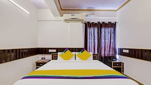A hotel room with a bed, two bedside tables, a nightstand, an air conditioning unit, and a window with curtains. - Itsy Hotels HSR Comfort (Bengaluru)