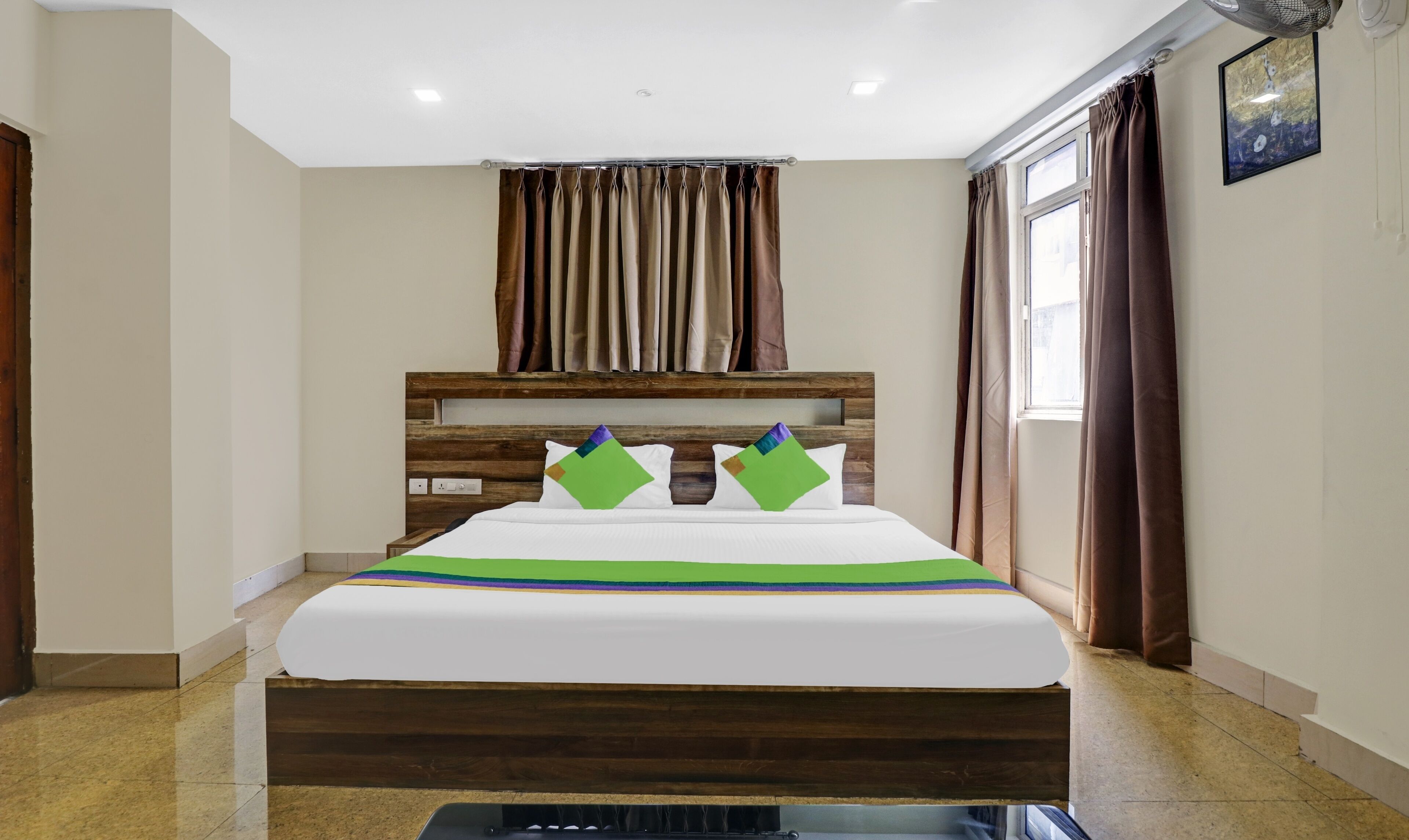 Ein Hotelzimmer mit einem Holzkopfteil, einem Bett mit einer grün-weiß gestreiften Bettdecke, zwei Kissen mit geometrischen Mustern, einem Deckenventilator und einem Fenster mit Vorhängen.
