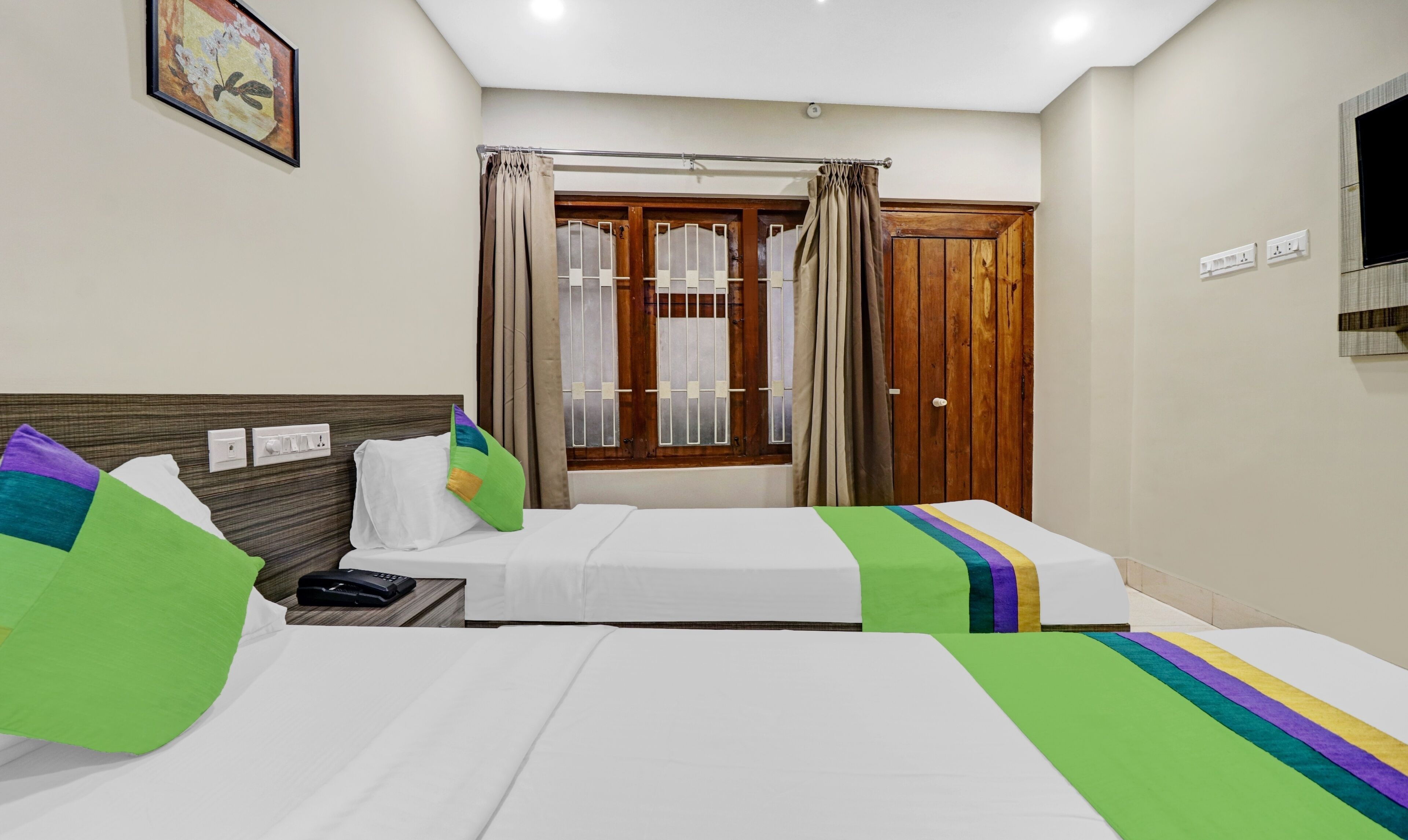 A hotel room with two single beds, a wooden headboard, a window with curtains, a wall-mounted TV, and a painting.