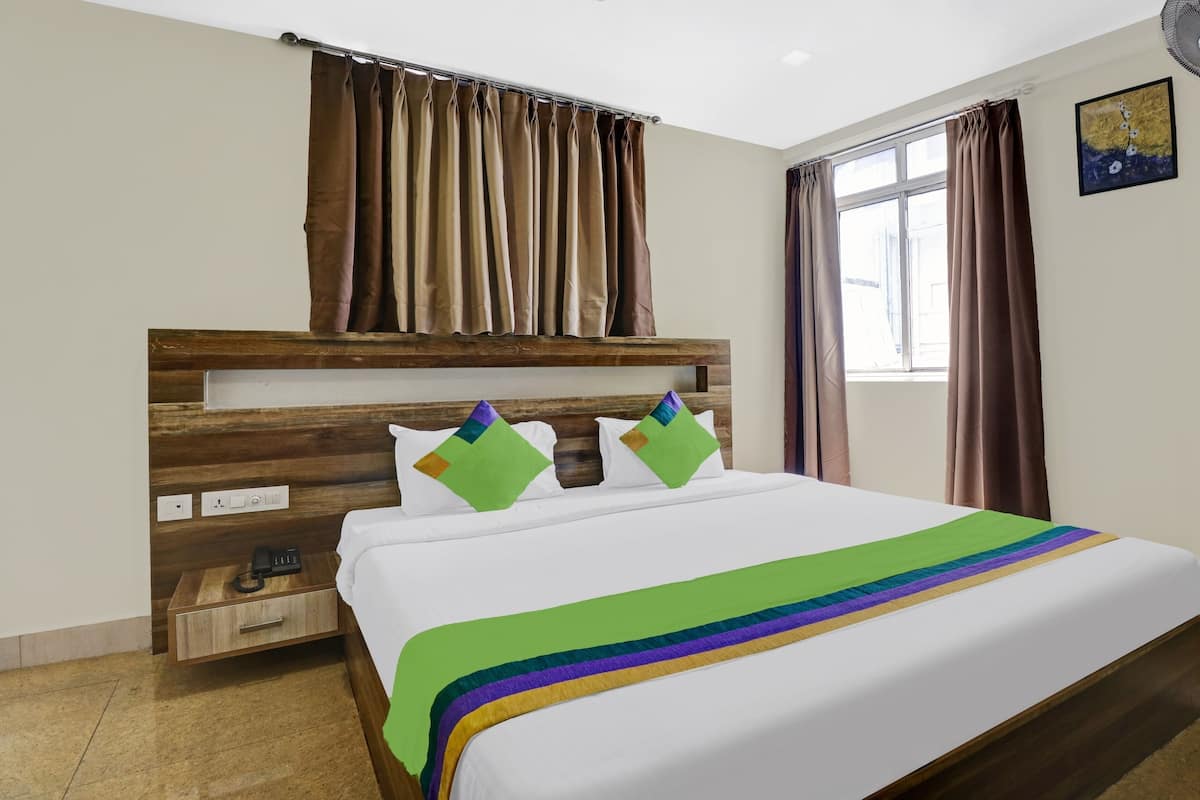 Una habitación de hotel con cabecera de madera, una cama con ropa de cama blanca y una colcha a rayas de arcoíris, una mesita de noche con un teléfono y una ventana con cortinas.