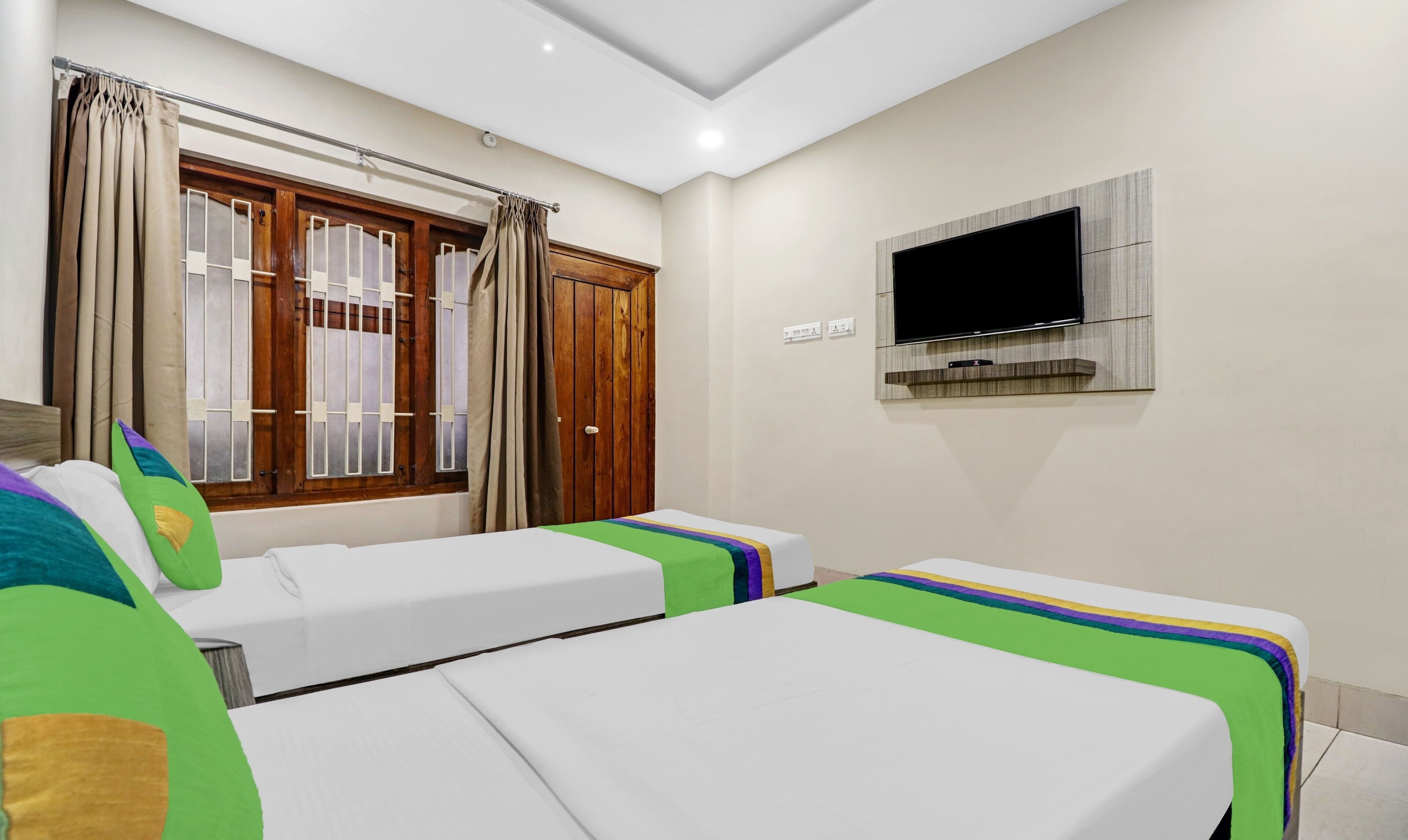A hotel room with two single beds, a TV mounted on the wall, and a window with curtains.
