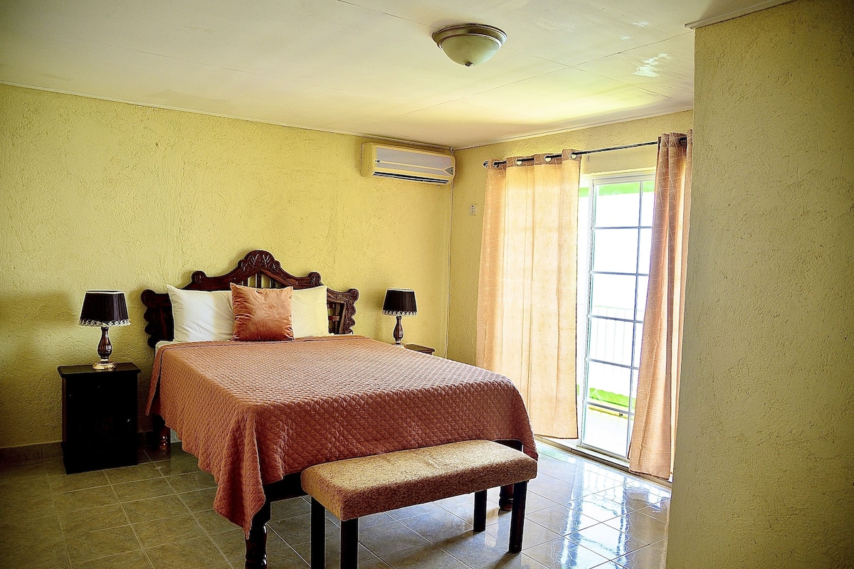 A bedroom with a bed, bedside tables, a bench, a window with curtains, and an air conditioning unit.