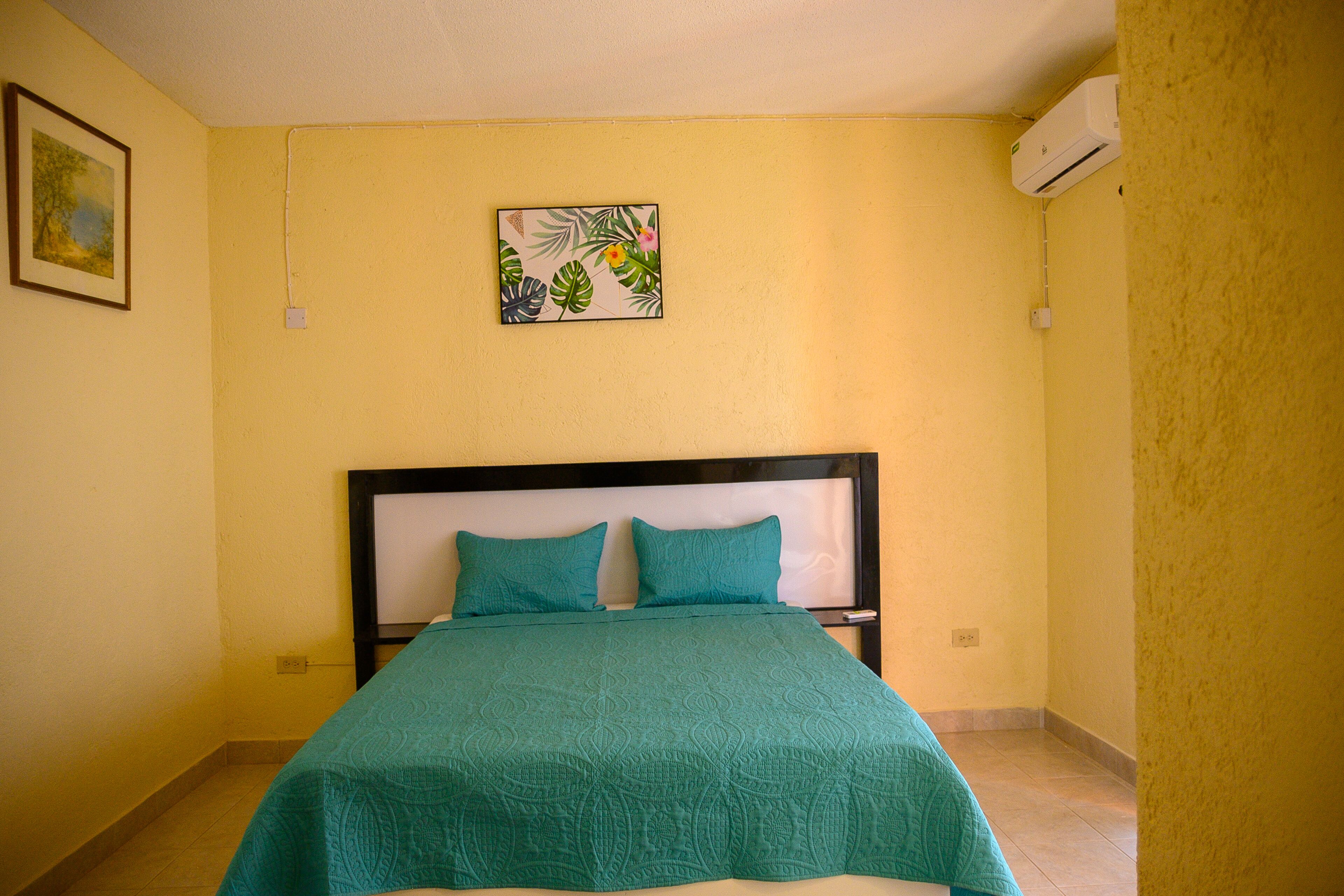 A bed with a teal bedspread and two pillows, a painting on the wall, and an air conditioning unit.
