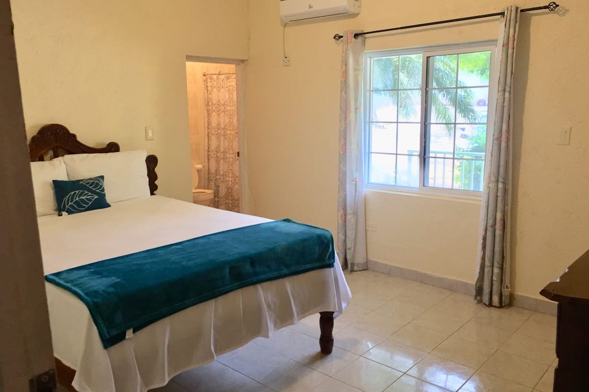 A bedroom with a bed, a window with curtains, a bathroom visible through an open door, and an air conditioning unit on the wall.